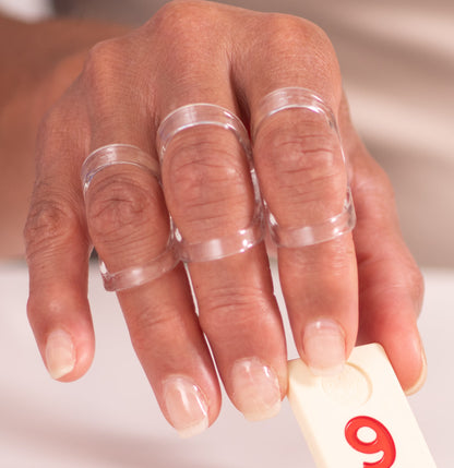 A person's hand is shown holding a domino with oval-8 clear finger splints on the fingers. No messy tape or straps; thin, lightweight - helps  manage Mallet Finger, limit Swan Neck or Hypermobility, prevent Trigger Finger/Thumb popping & stabilize Crooked, Jammed or Arthritic Fingers